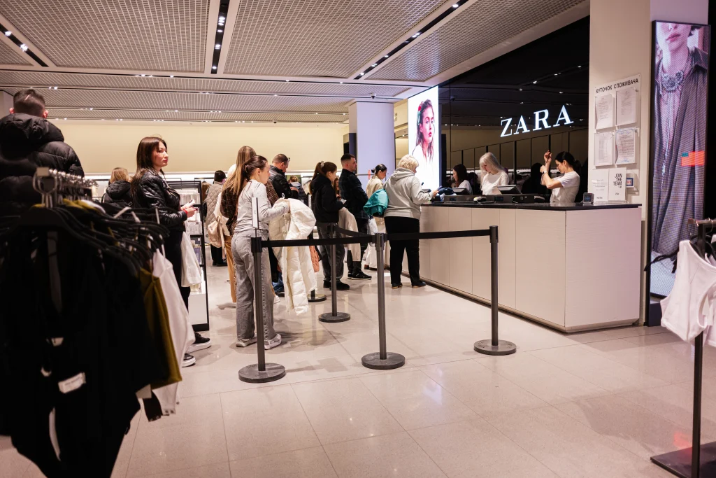 People standing in line at a Zara clothing store checkout counter. The store has bright lighting, a minimalist interior, and shopping racks with clothes visible in the foreground. Cashiers are assisting customers at the counter. Pessoas em fila no caixa de uma loja de roupas Zara. A loja tem iluminação forte, interior minimalista e araras de roupas visíveis no primeiro plano. As caixas estão atendendo os clientes no balcão.