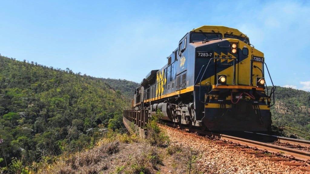 Locomotiva da MRS em pintura azul e amarela atravessa uma ferrovia em curva na serra, sob céu azul, cercada por mata atlântica. MRS freight locomotive in blue and yellow livery rounding a mountain curve, under a blue sky, surrounded by Atlantic Forest vegetation.