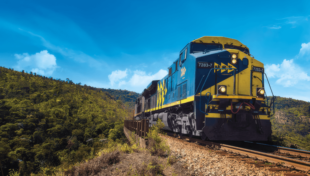 Locomotiva da MRS em pintura azul e amarela atravessa uma ferrovia em curva na serra, sob céu azul, cercada por mata atlântica. MRS freight locomotive in blue and yellow livery rounding a mountain curve, under a blue sky, surrounded by Atlantic Forest vegetation.