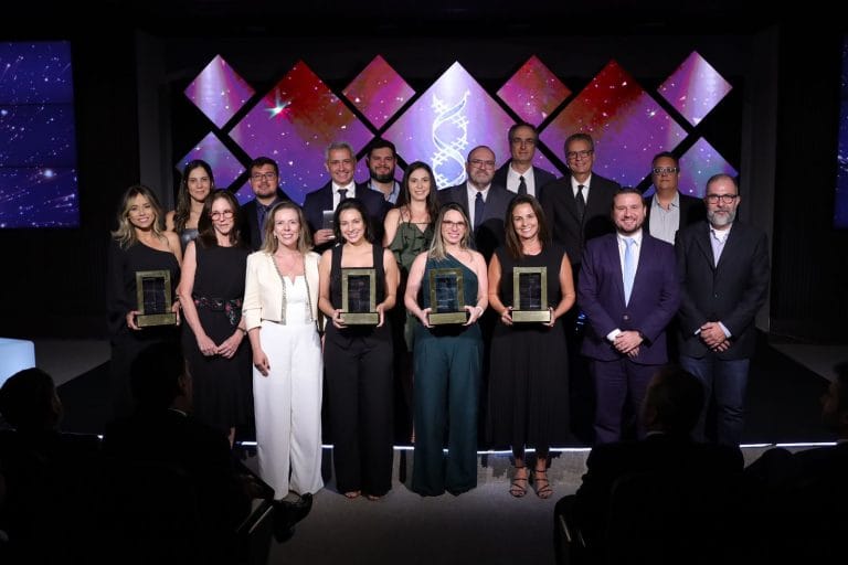 Cerimônia de premiação com grupo de pessoas no palco; quatro mulheres seguram troféus. Award ceremony with a group on stage; five women holding trophies.