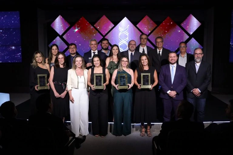 Cerimônia de premiação com grupo de pessoas no palco; quatro mulheres seguram troféus. Award ceremony with a group on stage; five women holding trophies.