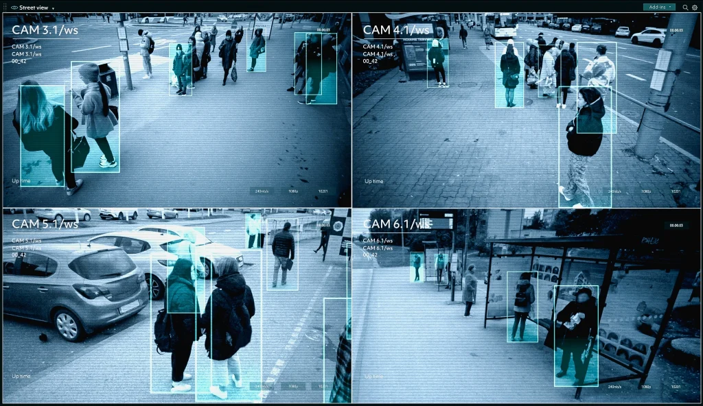 Imagem de videomonitoramento mostrando quatro telas com cenas urbanas, pessoas sendo detectadas e destacadas por caixas retangulares azuis nas imagens das câmeras de segurança.