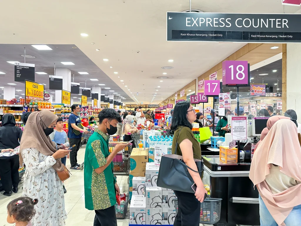 Área de checkout de supermercado movimentada com vários clientes aguardando em filas longas nos caixas numerados, ilustrando congestionamento e o risco de abandono de filas em ambiente varejista. Busy supermarket checkout area with multiple customers waiting in long lines at numbered checkout counters, illustrating the risk of queue abandonment in a retail environment.