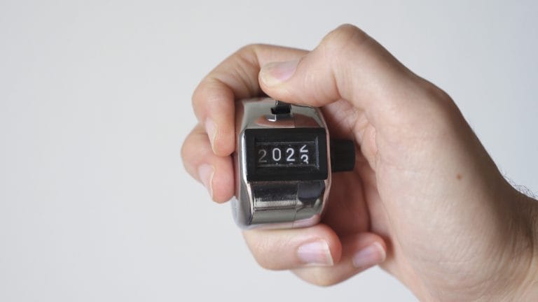 Uma fotografia em close-foco mostra uma mão segurando um contador manual metálico e prateado contra um fundo branco e liso. O polegar está posicionado sobre a alavanca de acionamento do dispositivo. O visor numérico do contador exibe os números "202", com o quarto dígito girando entre o "2" e o "3", sugerindo uma contagem em andamento.