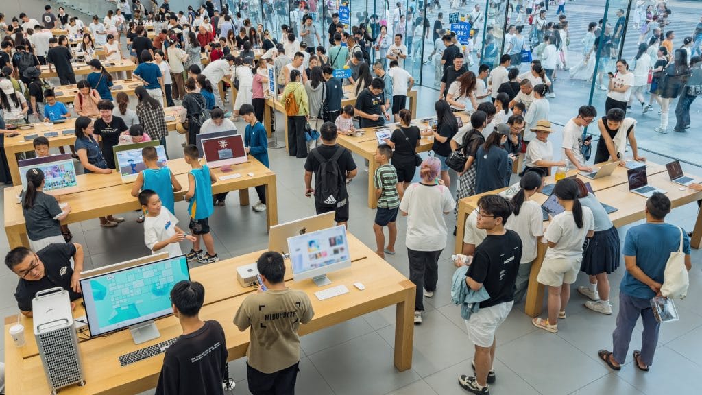 Uma fotografia aérea em ângulo picado mostra o interior de uma loja de tecnologia extremamente movimentada, com um design minimalista e moderno. Dezenas de pessoas circulam entre longas mesas de madeira clara, onde estão expostos computadores de mesa coloridos, notebooks e dispositivos móveis para teste. Os clientes, em sua maioria jovens, interagem com os produtos ou conversam entre si. tema: fluxo e conversão/traffic and conversion rate