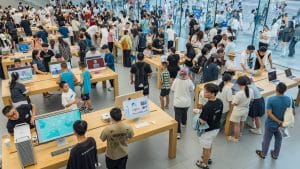Uma fotografia aérea em ângulo picado mostra o interior de uma loja de tecnologia extremamente movimentada, com um design minimalista e moderno. Dezenas de pessoas circulam entre longas mesas de madeira clara, onde estão expostos computadores de mesa coloridos, notebooks e dispositivos móveis para teste. Os clientes, em sua maioria jovens, interagem com os produtos ou conversam entre si. tema: fluxo e conversão/traffic and conversion rate