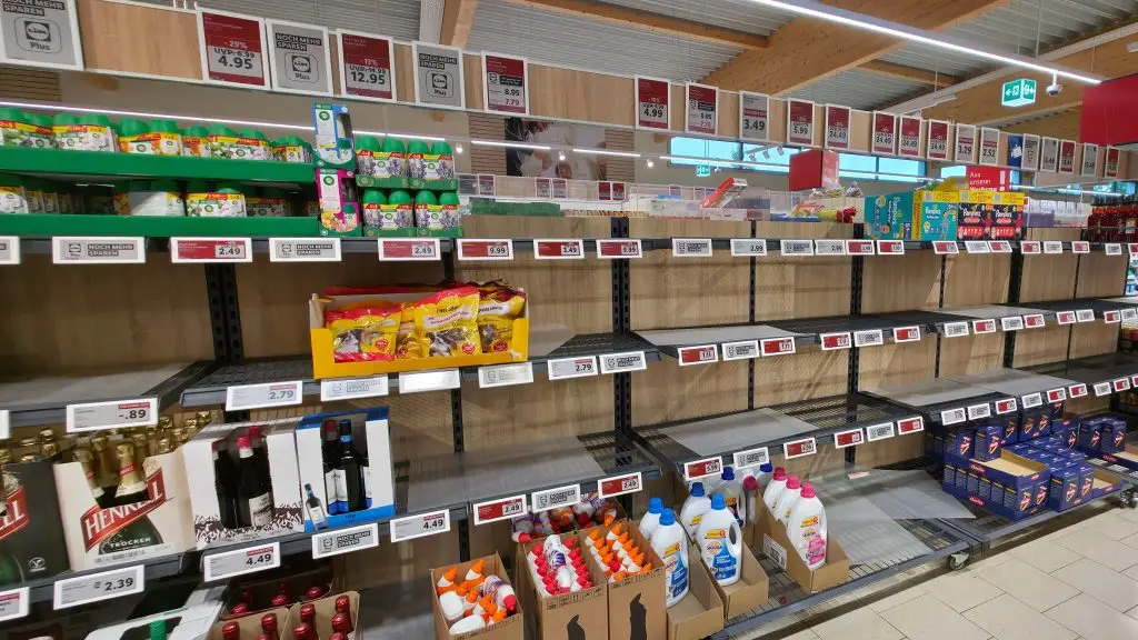 Ilustração do conceito de ruptura operacional (operational stockout). Uma fotografia de um corredor de prateleiras em um supermercado. O topo exibe plantas e flores com cartazes suspensos de preços promocionais. As prateleiras de metal abaixo contêm sacos de ração para animais de estimação, garrafas de vinho frisante e várias prateleiras vazias. As prateleiras inferiores mostram caixas de varejo com garrafas de detergente líquido e amaciante de roupas. Várias etiquetas de preço vermelhas e brancas são visíveis. A perspectiva mostra o corredor se estendendo ao fundo com mais prateleiras e sinalização de teto.