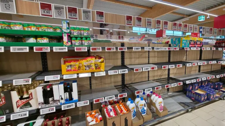 Ilustração do conceito de ruptura operacional (operational stockout). Uma fotografia de um corredor de prateleiras em um supermercado. O topo exibe plantas e flores com cartazes suspensos de preços promocionais. As prateleiras de metal abaixo contêm sacos de ração para animais de estimação, garrafas de vinho frisante e várias prateleiras vazias. As prateleiras inferiores mostram caixas de varejo com garrafas de detergente líquido e amaciante de roupas. Várias etiquetas de preço vermelhas e brancas são visíveis. A perspectiva mostra o corredor se estendendo ao fundo com mais prateleiras e sinalização de teto.
