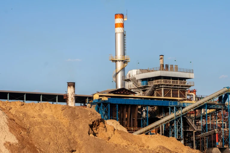 Vista do pátio de uma usina de bioenergia mostrando uma grande pilha irregular de biomassa a granel em primeiro plano. Ao fundo, a infraestrutura industrial exibe estruturas metálicas, esteiras transportadoras azuis e uma alta chaminé cinza com listras laranjas e brancas no topo, sob um céu azul. View of a bioenergy plant yard featuring a large, irregular bulk biomass stockpile in the foreground. In the background, the industrial facility infrastructure shows steel structures, blue conveyor belts, and a tall grey smokestack with orange and white stripes at the top under a clear blue sky. Conceito: Volumetria / Volumetric Measurement